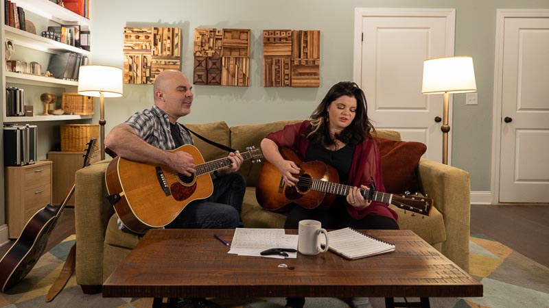 Will Robertson and Rebecca Loebe playing guitar and singing on a couch behind a coffee table with sheet music on it